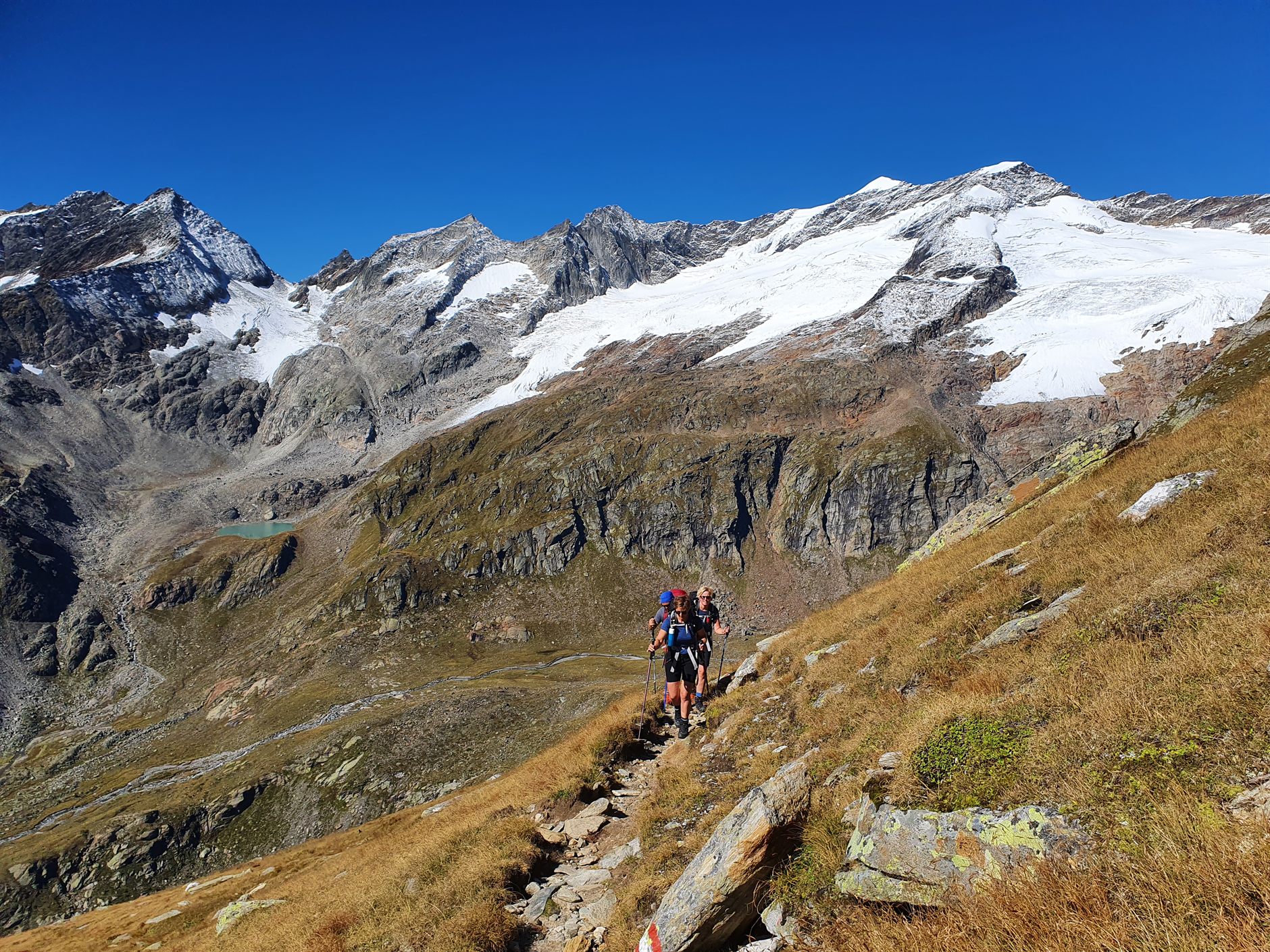Wandelaars op een bergpad hoog boven een dal met besneeuwde toppen en gletsjers